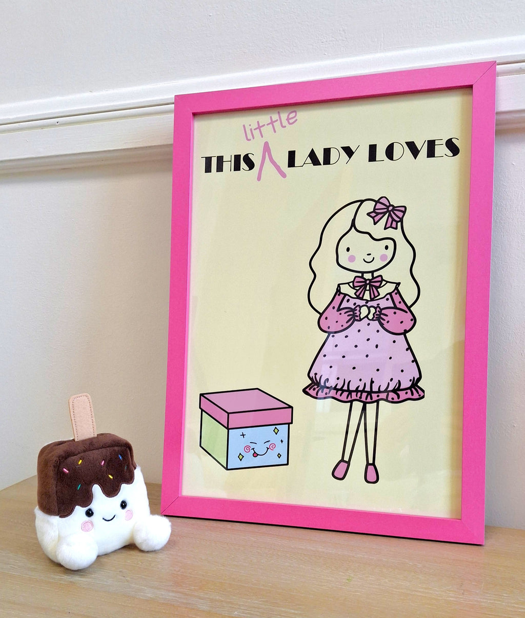 A framed art print with a bright pink frame, featuring 'THIS LITTLE LADY LOVES' branding. The frame is displayed next to a small plush ice cream toy.