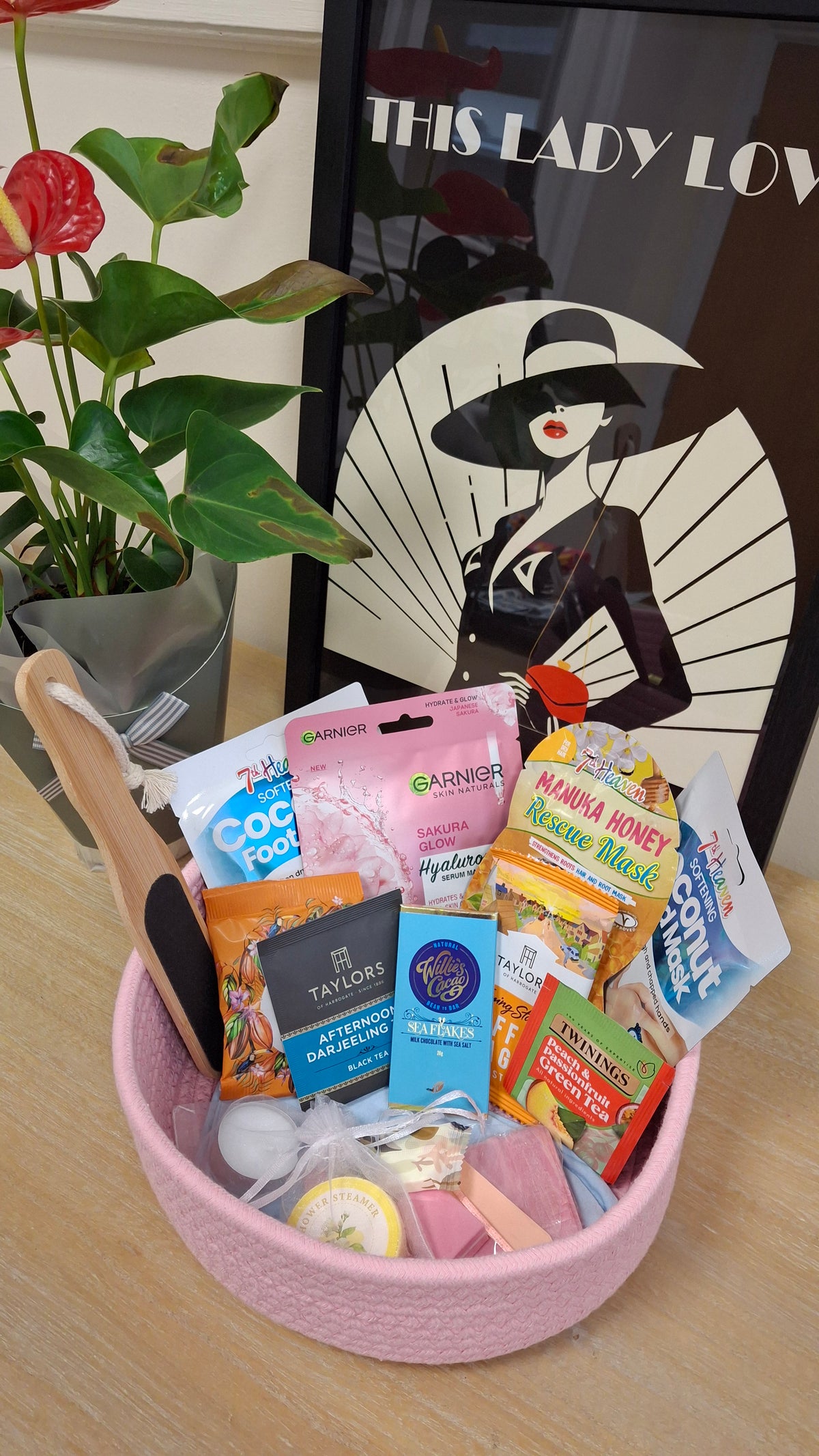 A pink woven basket containing the content of the pamper box with This Lady Loves branding poster and a pot plant.
