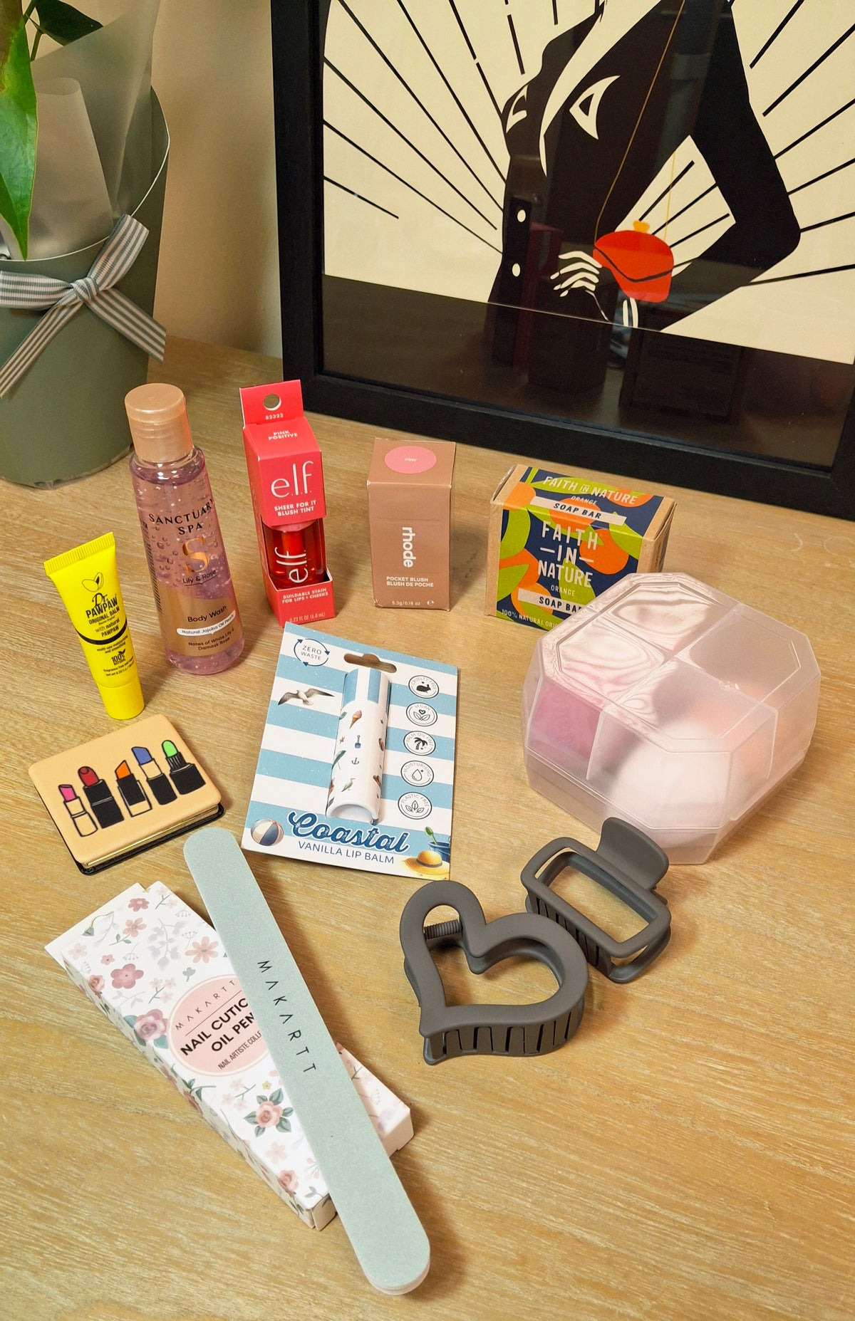 A flat-lay arrangement of various beauty and personal care items on a light wood surface, including pink beauty blenders , hair clips, a nail buffer, lip gloss and other cosmetic products.