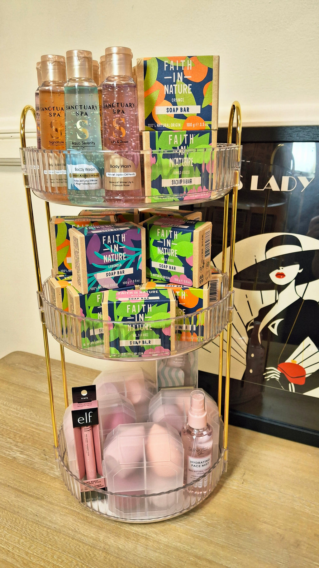 A clear acrylic and gold metal cosmetic organizer filled with various beauty products, including small bottles, colorful boxes, and pink makeup sponges, set on a light wood floor in front of a framed print.
