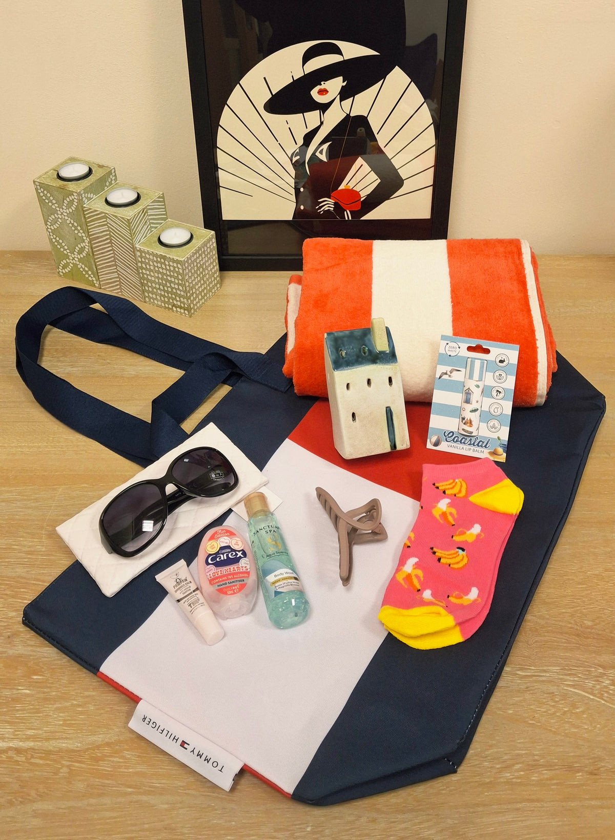 A curated gift set arranged on a light wood tabletop in front of a framed art print. The set includes a Tommy Hilfiger beach bag in white, red and navy, a red and white striped beach towel, a pair of trainer socks, sunglasses and pouch, hand sanitizer, lip balm, shower gel, Dr Paw Paw balm, Coastal House tealight holder and hair clip.