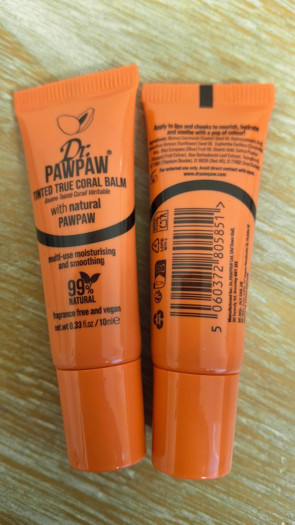 Two orange Dr. Pawpaw lip balm tubes standing upright on a light wood surface, showing the front label on the left and the back with ingredients list.