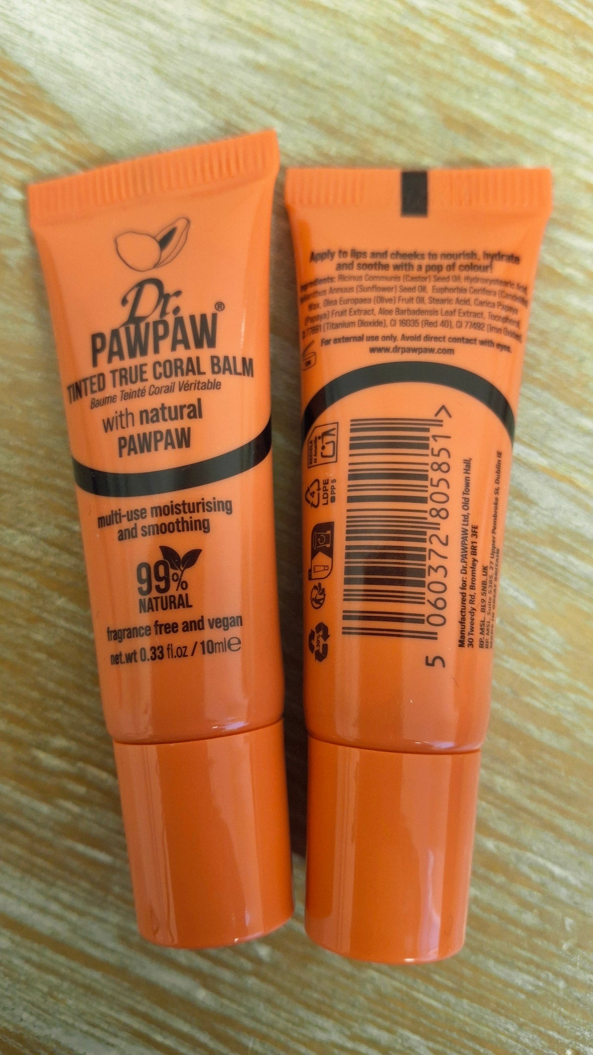 Two orange Dr. Pawpaw lip balm tubes standing upright on a light wood surface, showing the front label on the left and the back with ingredients list.
