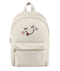 A cream-colored nylon backpack with a gold zipper and a cute embroidered face on the front pocket showing a winking eye, pink cheeks, and a sticking-out tongue.