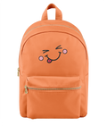 An orange nylon backpack with a cute embroidered face on the front pocket, featuring a winking eye, pink cheeks, and a sticking-out tongue. The backpack has a top handle, a main zippered compartment, and a front zippered pocket with gold-tone zippers.