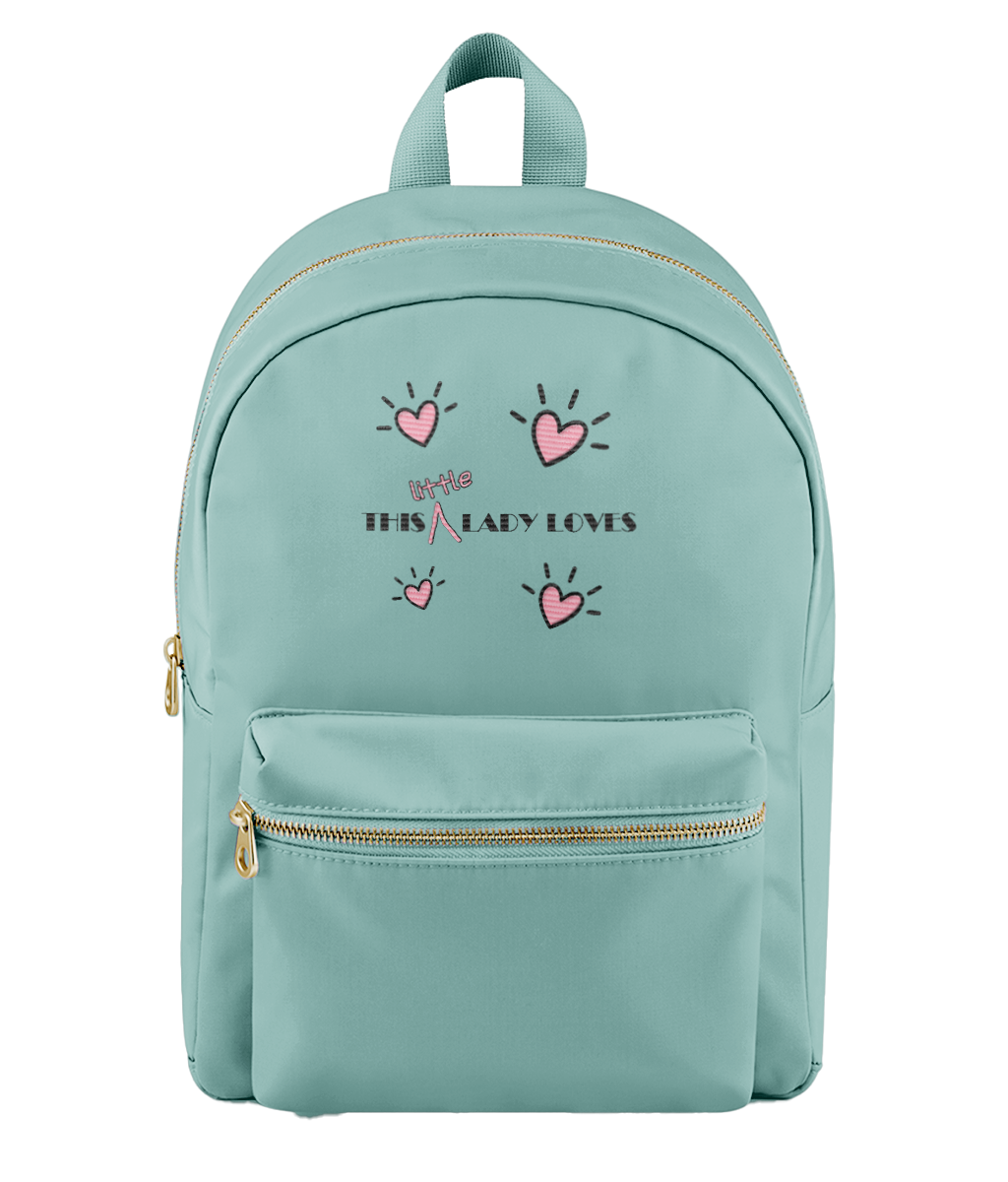 A mint green backpack with a front pocket, gold-tone zippers, and a graphic print featuring pink hearts and the text 'This Little Lady Loves' on the front, set against a plain white background.