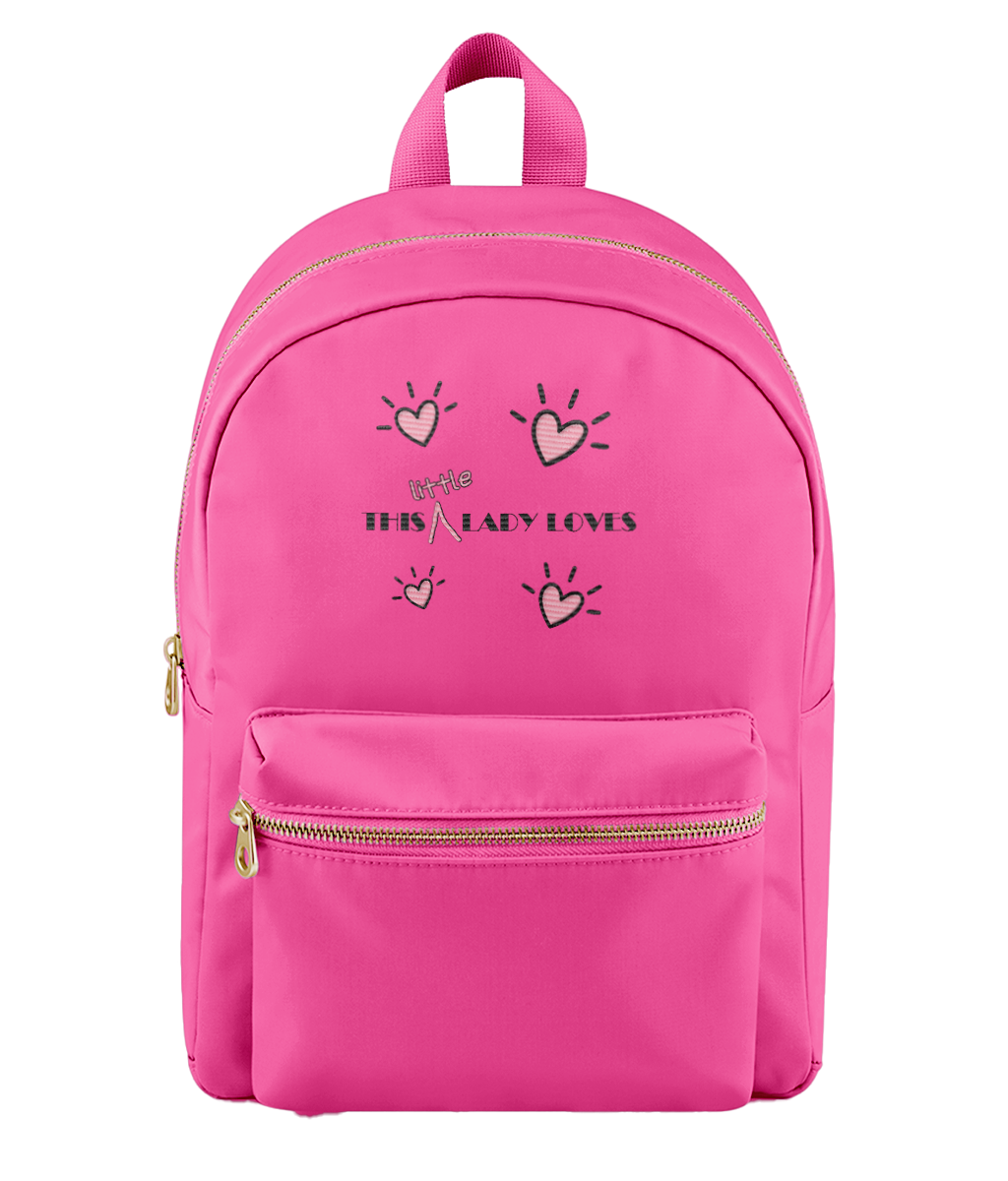 A bright pink nylon backpack with a front zippered pocket and top handle, featuring a graphic print of hand-drawn hearts and the text 'THIS LITTLE LADY LOVES' in black with a small pink bow.